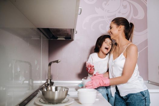 housewife mom in pink gloves washes dishes with her son by hand in the sink with detergent. A girl in white and a child with a cast cleans the house and washes dishes in homemade pink gloves.A child with a cast washes dishes and smiles.