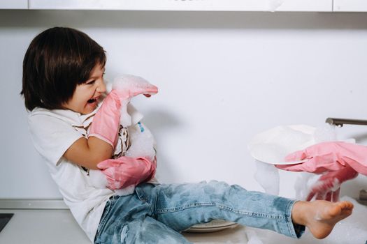 housewife mom in pink gloves washes dishes with her son by hand in the sink with detergent. A girl in white and a child with a cast cleans the house and washes dishes in homemade pink gloves.A child with a cast washes dishes and smiles.