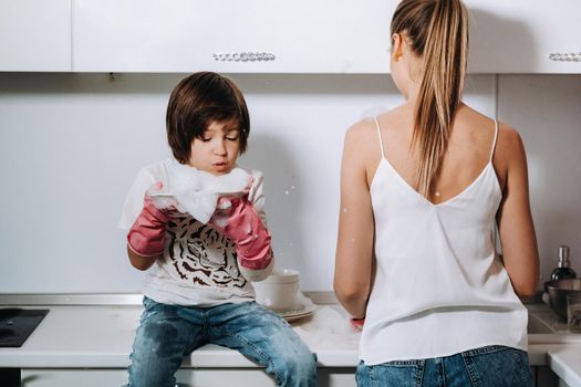 housewife mom in pink gloves washes dishes with her son by hand in the sink with detergent. A girl in white and a child with a cast cleans the house and washes dishes in homemade pink gloves.A child with a cast washes dishes and smiles.