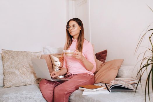 a girl in the morning in pajamas at home working on a laptop with drinking coffee, a girl self-isolated at home and resting on the couch and watching a laptop.Household chores.