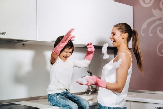 housewife mom in pink gloves washes dishes with her son by hand in the sink with detergent. A girl in white and a child with a cast cleans the house and washes dishes in homemade pink gloves.A child with a cast washes dishes and smiles.