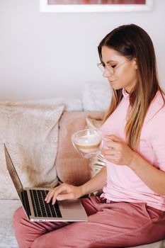 a girl in the morning in pajamas at home working on a laptop with drinking coffee, a girl self-isolated at home and resting on the couch and watching a laptop.Household chores.