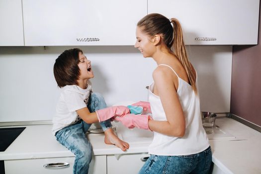 housewife mom in pink gloves washes dishes with her son by hand in the sink with detergent. A girl in white and a child with a cast cleans the house and washes dishes in homemade pink gloves.A child with a cast washes dishes and smiles.