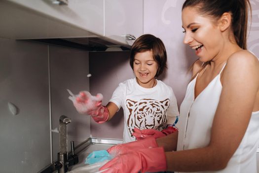 housewife mom in pink gloves washes dishes with her son by hand in the sink with detergent. A girl in white and a child with a cast cleans the house and washes dishes in homemade pink gloves.A child with a cast washes dishes and smiles.