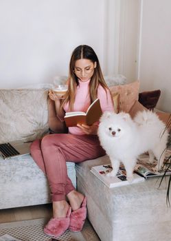 a girl in pajamas at home reads a book with her dog Spitzer, the Dog and its owner are resting on the sofa and reading a book.Household chores.