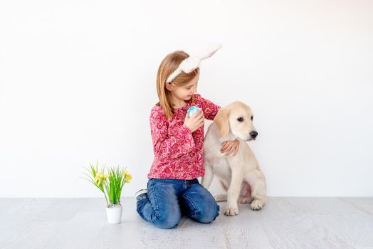 Funny little girl in bunny ears holding colored egg and hugging young dog in light interior