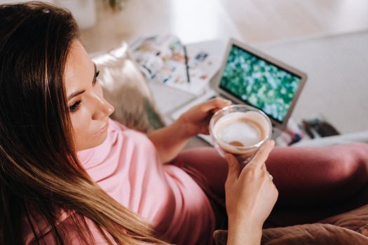 a girl in the morning in pajamas at home working on a laptop with drinking coffee, a girl self-isolated at home and resting on the couch and watching a laptop.Household chores.