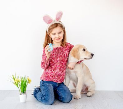 Funny little girl in bunny ears holding colored egg and hugging young dog in light interior