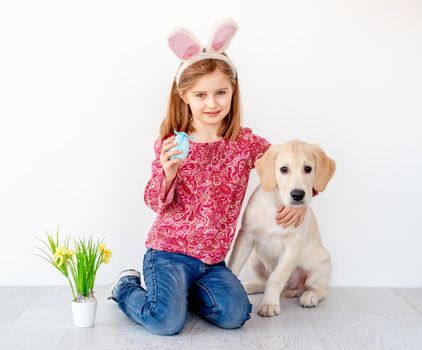 Funny little girl in bunny ears holding colored egg and hugging young dog in light interior