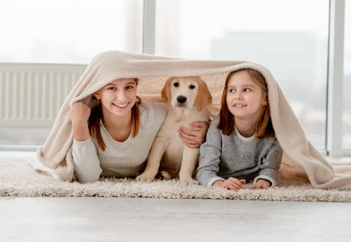 Lovely sisters and golden retriever puppy under plaid on floor in room