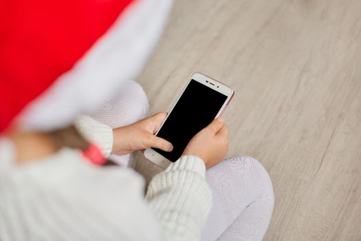 Faceless child holding phone with blank screen while posing backwards, wearing sweater and santa hat, sitting on floor, using mobile phone for video calling or playing games.