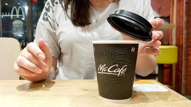 Ukraine, Kiev - September 09, 2019: A girl covers a black plastic paper lid with a hot black coffee maker in McDonalds, McCafe sitting at a table in a fast food restaurant.