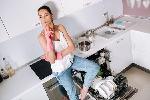 a housewife girl in pink gloves after cleaning the house sits tired in the kitchen.In the white kitchen, the girl has washed the dishes and is resting.Lots of washed dishes.
