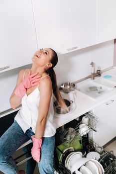 housewife girl in pink gloves after cleaning the house is emotional and tired in the white kitchen, the girl has washed the dishes and is resting and expressing emotions.A lot of washed dishes.