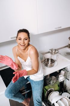 housewife girl in pink gloves after cleaning the house is emotional and tired in the white kitchen, the girl has washed the dishes and is resting and expressing emotions.A lot of washed dishes.