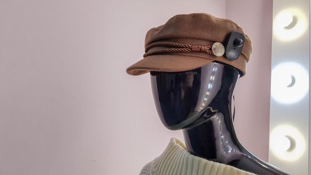 Portrait of a black mannequin with a brown hat in a women fashion store. Mannequin woman in a hat on a pink wall background and a large white mirror with backlight and light bulbs.