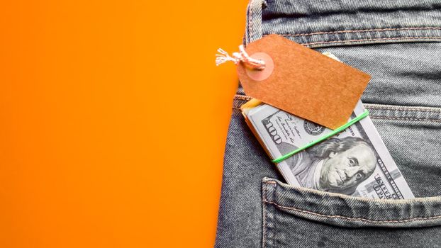 Many hundreds of dollars banknotes in a jeans pocket in a purse with a cardboard tag, price tag. Wallet and dollars in jeans pocket. Copy space, place for text, flat lay. Background, holiday concept. Black Friday - International Day of Shopping, Promotions, Discounts, Sales. Sales season is the fourth Thursday of November. Blue jeans on a colored background.