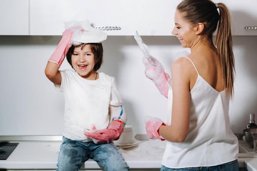 housewife mom in pink gloves washes dishes with her son by hand in the sink with detergent. A girl in white and a child with a cast cleans the house and washes dishes in homemade pink gloves.A child with a cast washes dishes and smiles.