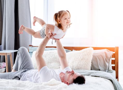 Father playing with little daughter in the bed and holding her in the air in his hands. Young dad with child in the morning time in sunny room. Happy family moments together