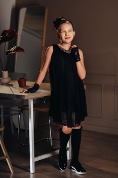 A stylish little girl in a black dress stands in the interior near the table.