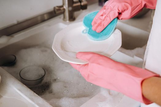 housewife girl in pink gloves washes dishes by hand in the sink with detergent. The girl cleans the house and washes dishes in gloves at home