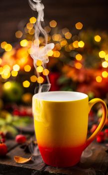 a red and yellow mug with a steaming drink. Fruits and spices are all around on a wooden table. lights of garlands are burning behind