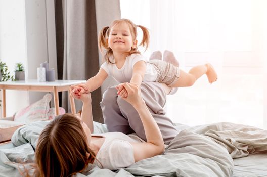Young mother playing with daughter in the bed swinging cute child on her legs. Caucasian mom and her kid have fun in the bedroom with sunlight. Beautiful family morning time