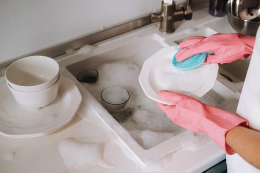 housewife girl in pink gloves washes dishes by hand in the sink with detergent. The girl cleans the house and washes dishes in gloves at home