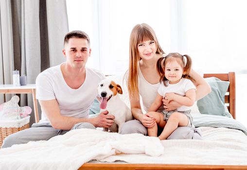 Lovely portrait of family morning in the bedroom. Beautiful parents with their daughter staying in the bed with dog and looking at the camera. Mother, father and child wearing pajamas with pet