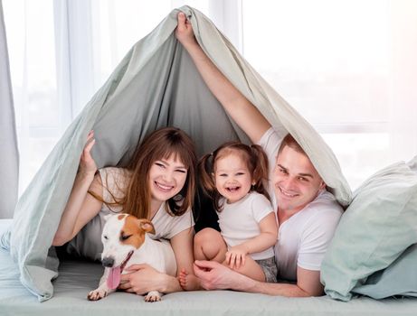 Smiling family with dog under the blanket in the bed. Beautiful morning moments of mother, father and little daughter with pet under coverlet. Portrait of happy family