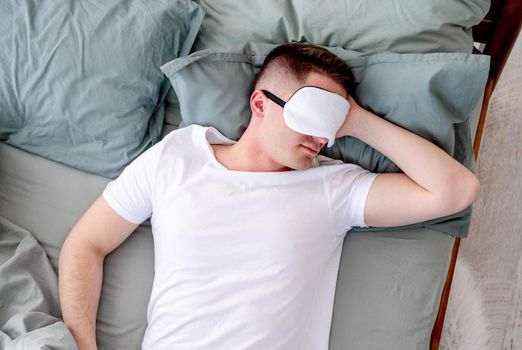 Young man sleeping in the bed wearing white t-shirt and eye mask. Guy lying on his back and napping in the room with sunlight in the morning. Concept of resting time