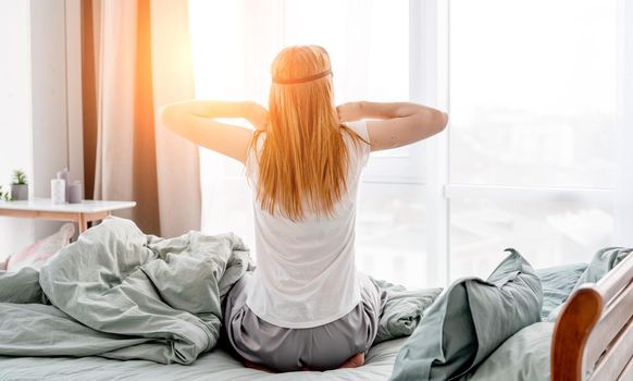 Attractive girl wakes up in sunny room with panoramic window and stretch her body in the bed. Back view of young woman wearing pajama in the morning time in the bedroom. Concept of good rest and relax