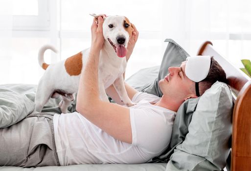Man playing with dog in the bed in the morning time. Young guy wakes up and doggy pet standing on him and looking at the camera. Concept of friendship beetween human and animal