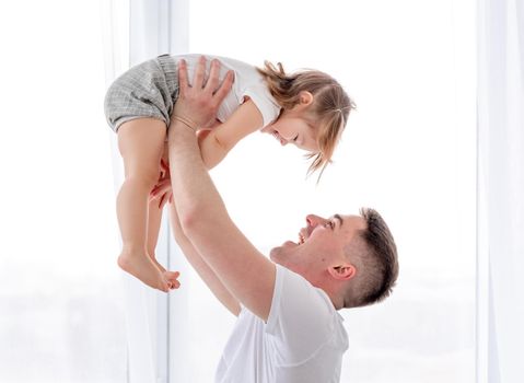 Young father spending morning with his little daughter holding her in his hands against the window. Smiling child with her parent in sunny room. Beautiful moments of dad with his kid in the bedroom