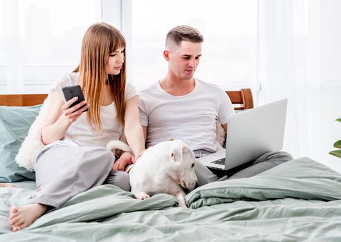 Young couple in the bed with gadgets and cute dog. Beautiful woman and man spending time together in the bedroom and looking at laptop and smartphone. Family morning with pet