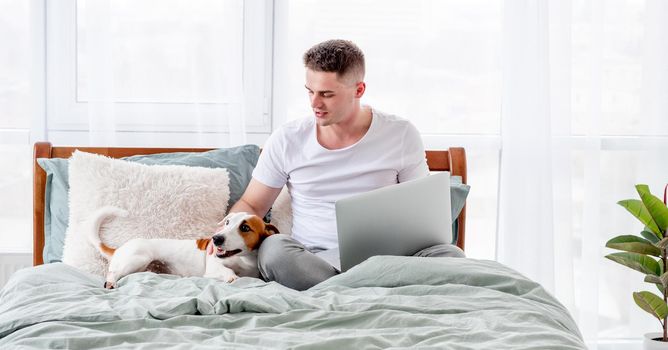 Young handsome man sitting in the bed with holding laptop on his legs and petting cute dog. Guy working from home in the bedroom and his doggy pet staying close to him. Concept of freelance