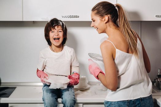 housewife mom in pink gloves washes dishes with her son by hand in the sink with detergent. A girl in white and a child with a cast cleans the house and washes dishes in homemade pink gloves.A child with a cast washes dishes and smiles.