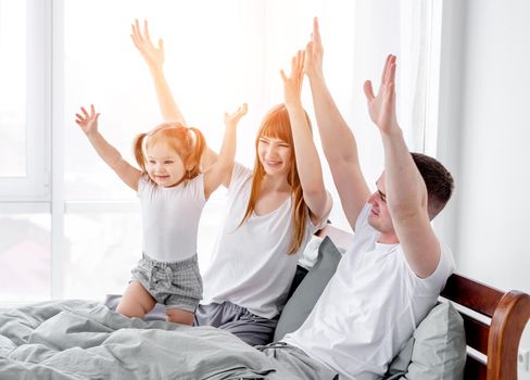 Sunny family mornents in the bedroom. Beautiful parents with their daughter staying in the bed and enjoying time together. Mother, father and child holding hands up and smiling