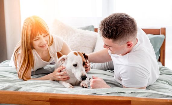 Lovely portrait of young couple in the bed with cute dog. Family morning moments. Attractive girl and guy in the bedroom petting their doggy and smiling. Young wife and husband with pet