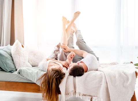 Beautiful young couple lying in the bed with legs up in sunny room with panoramic window. Girl and guy wearing pajamas spend time together in the bedroom. Lovely family morning