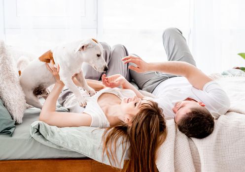 Beautiful family with cute dog in the bed in the room with sunlight. Concept of happy family and love. Young woman and man with their pet in morning time