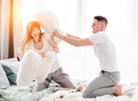 Beautiful young couple fighting with pillows in the bed and smiling. Girl and guy have fun in the bedroom with sunlight and laughing. Lovely family moments and enjoying of life