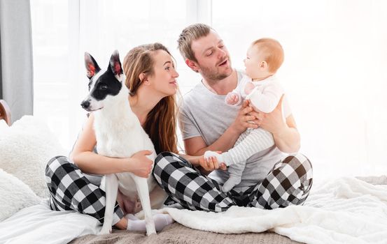 Family with baby boy sitting on the bed and hugging cute dog. Mother and father holding their son on legs in sunny room. Beautiful parenthood time. Pet with owners