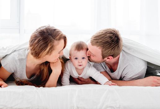 Mother and father lying in the bed under blanket with their son and kissing baby on his cheeks. Beautiful family morning together. Happy parenthood