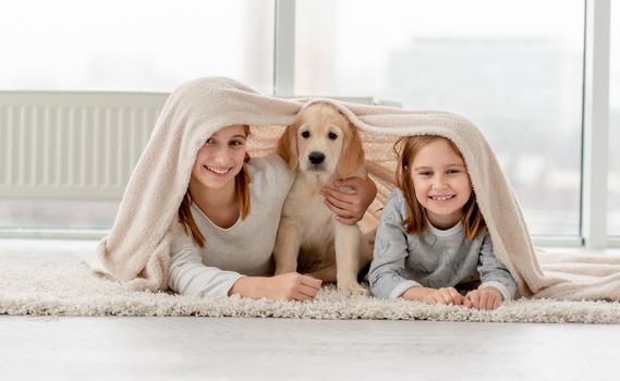 Lovely sisters and golden retriever puppy under plaid on floor in room