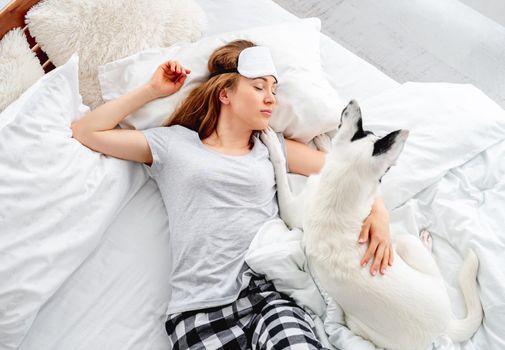 Beautiful long hair girl lying in the bed on her back with cute dog and sleeping. Pretty young woman resting with pet. Female person wearing eye sleeping mask napping with doggy