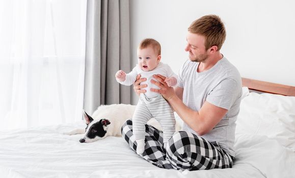 Young father sitting in the bed with little son and cute dog in the room with sunlight. Man staying in the bedroom with his child and pet. Guy with kid and doggy in the morning time