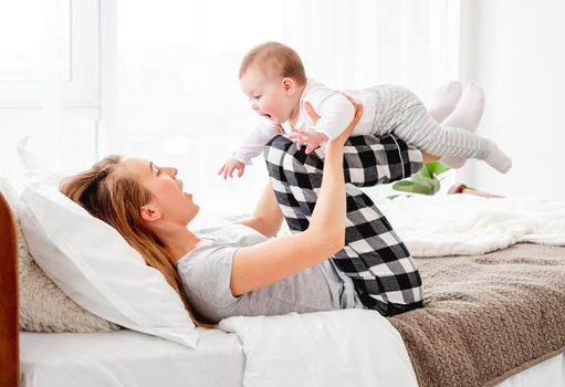 Beautiful young mother holding little baby boy on her legs and looking at the child. Pretty girl playing with son kid in the bed in morning time. Happy family moments