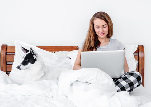 Beautiful girl wearing pajamas sitting in the bed with laptop working and cute doggy lying close to her. Young woman freelancer at home with pet in morning time typing on notebook