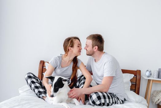 Beautiful caucasian couple sitting in the bed with cute dog. Young man and woman kissing each other anf pet lying close to them. Portrait of happy lovely family with doggy